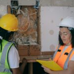 Two workers in protective gear inspecting and taking notes during a construction site electrical inspection.